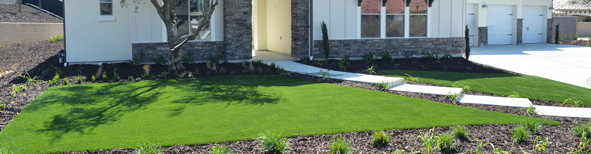 Artificial turf front yard installation on a modern farmhouse home with stone accents, mulch flower beds, young olive tree, and river rock drought-tolerant landscaping